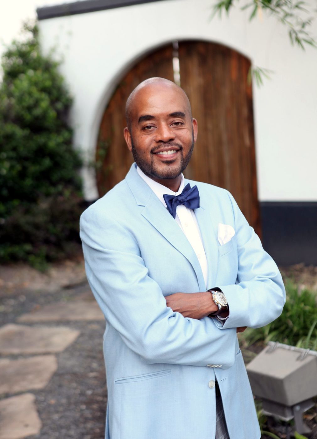 Jones Mays in a light blue blazer and navy bow tie, arms crossed, in front of an arched wooden door